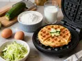 Gaufre courgette dorée dans un gaufrier, entourée d'ingrédients : courgettes râpées, œufs, farine et lait.