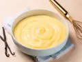Un bol de crème pâtissière onctueuse, parsemée de points de vanille, avec des gousses de vanille et un fouet doré à proximité.
