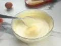 Préparation d'un tiramisu : une cuillère de pâte à bombe aérienne est prélevée d'un bol en verre. Un œuf et des biscuits à la cuillère sont visibles.