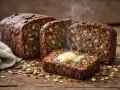 Tranche de pain nordique recette, garnie de beurre fondant et de vapeur s'échappant. Des graines et des flocons d'avoine parsèment la planche en bois.