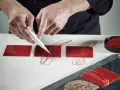 Mains d'un pâtissier créant un décor royal chocolat sur des carrés rouges brillants.
