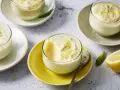 Plusieurs verrines de mousse au citron, garnies de zestes, sur des assiettes claires et jaunes. Un citron coupé est visible à droite.