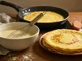 Préparation d'une recette de crêpes : pâte dans un bol, crêpes cuites dans une poêle et empilées, œufs et farine.