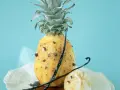 Un ananas rôti, parsemé de clous de girofle et de gousses de vanille, sur un fond bleu pâle. Un délice sucré et épicé.