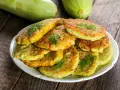 Pile de beignets de courgette dorés et moelleux, garnis de brins d'aneth frais, sur une assiette blanche. Deux courgettes vertes floues en arrière-plan.