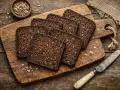 Tranches de pumpernickel sur une planche en bois, prêtes pour une délicieuse recette. Un bol de graines et un couteau complètent la scène rustique.