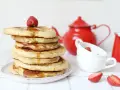 Pile de pancake sans gluten, nappés de sirop et garnis d'une fraise. Une théière rouge et des tasses complètent la scène.