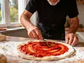 Un homme prépare une pizza, étalant un coulis de tomates sur la pâte avec une cuillère. La farine est saupoudrée sur la table.