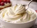 Une cuillère prélève une portion de chantilly mascarpone onctueuse dans un bol blanc. Des fruits rouges sont visibles en arrière-plan.