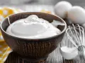Un bol rempli de crème fouettée, blanc en neige, prêt à être utilisé pour une pâtisserie. Des œufs et un fouet sont visibles à proximité.