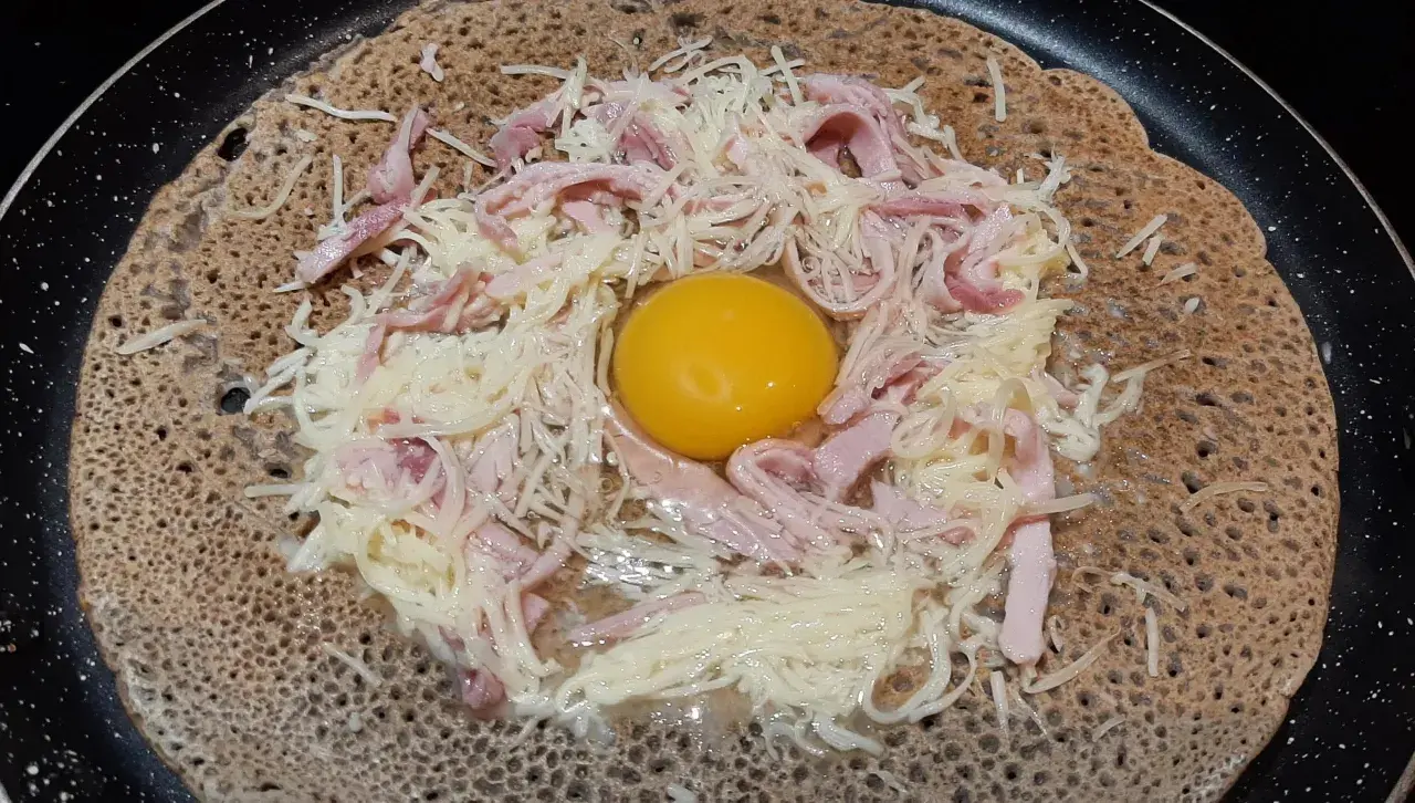Une galette sarrasin garnie d'un œuf, de jambon et de fromage râpé, prête à être cuite dans une poêle.