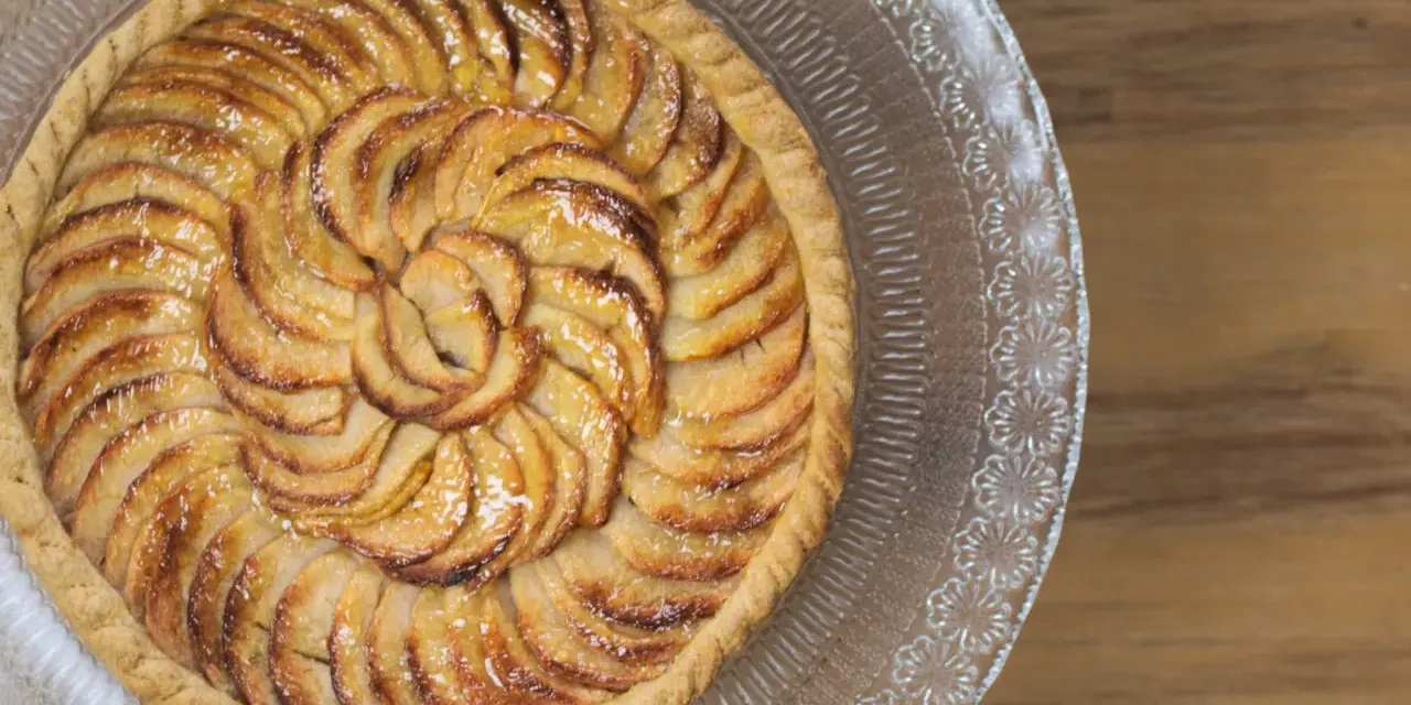 Un magnifique appareil tarte aux pommes, aux pommes caramélisées disposées en spirale sur une pâte dorée, sur un plat en verre décoré.