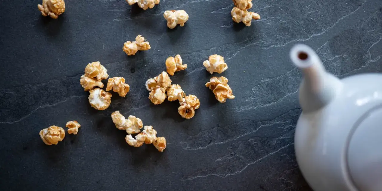 Pop corn caramel éparpillé sur une surface sombre, avec une théière blanche en arrière-plan.
