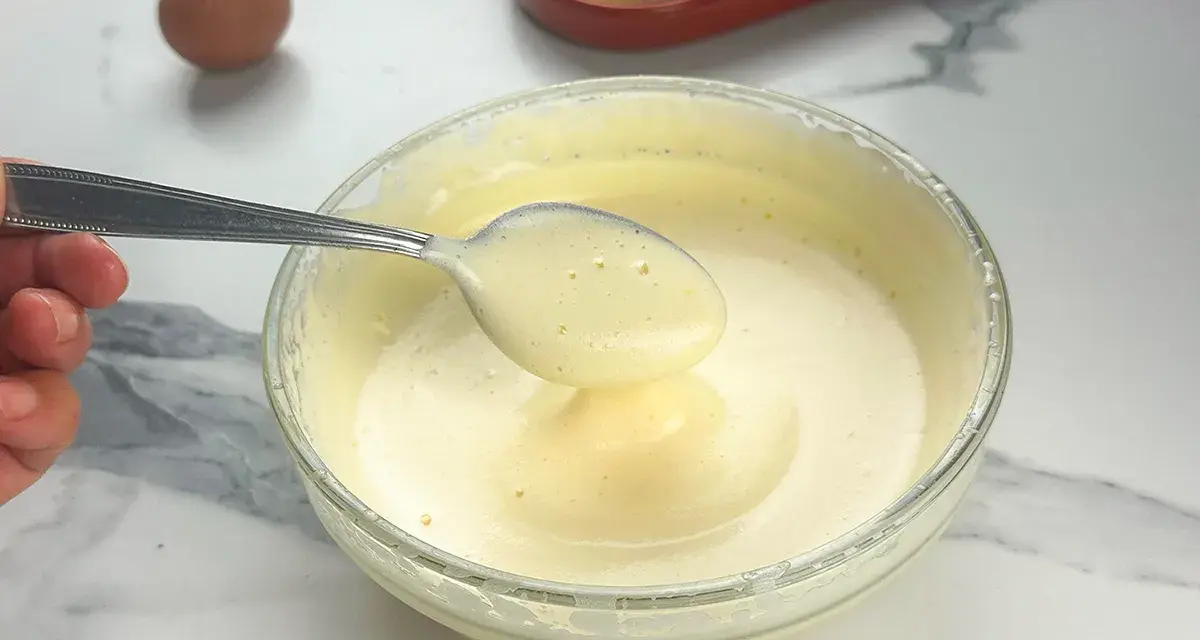 Préparation d'un tiramisu : une cuillère de pâte à bombe aérienne est prélevée d'un bol en verre. Un œuf et des biscuits à la cuillère sont visibles.