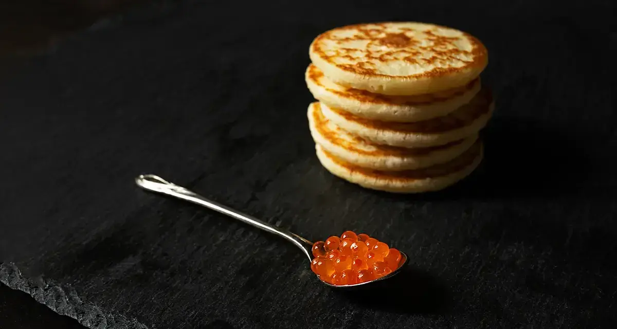 Une cuillère de caviar rouge sur une assiette noire, à côté d'une pile de blinis dorés. Parfait pour une recette blinis gourmande.