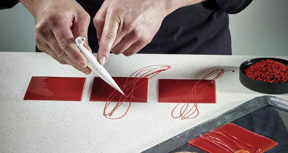 Mains d'un pâtissier créant un décor royal chocolat sur des carrés rouges brillants.