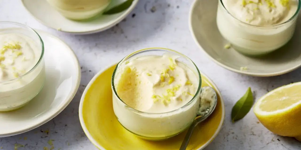 Plusieurs verrines de mousse au citron, garnies de zestes, sur des assiettes claires et jaunes. Un citron coupé est visible à droite.