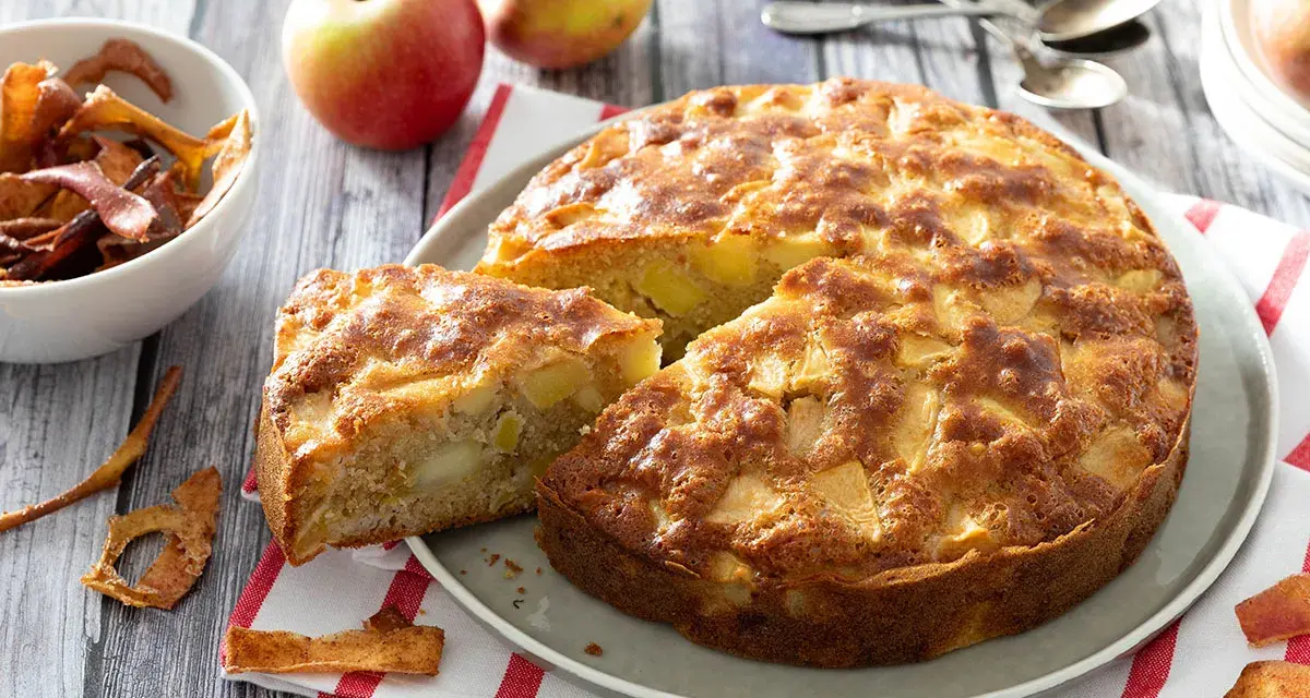 Un délicieux gâteau aux pommes, doré et moelleux, avec des morceaux de fruits visibles. Des pommes fraîches et des chips de pommes complètent la scène.