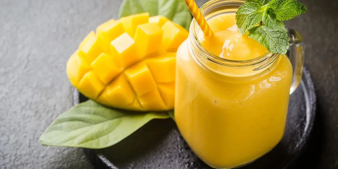 Un smoothie mangue onctueux dans un pot en verre, garni de menthe fraîche, à côté d'une moitié de mangue coupée en dés.