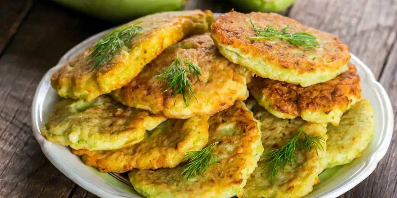 Pile de beignets de courgette dorés et moelleux, garnis de brins d'aneth frais, sur une assiette blanche. Deux courgettes vertes floues en arrière-plan.