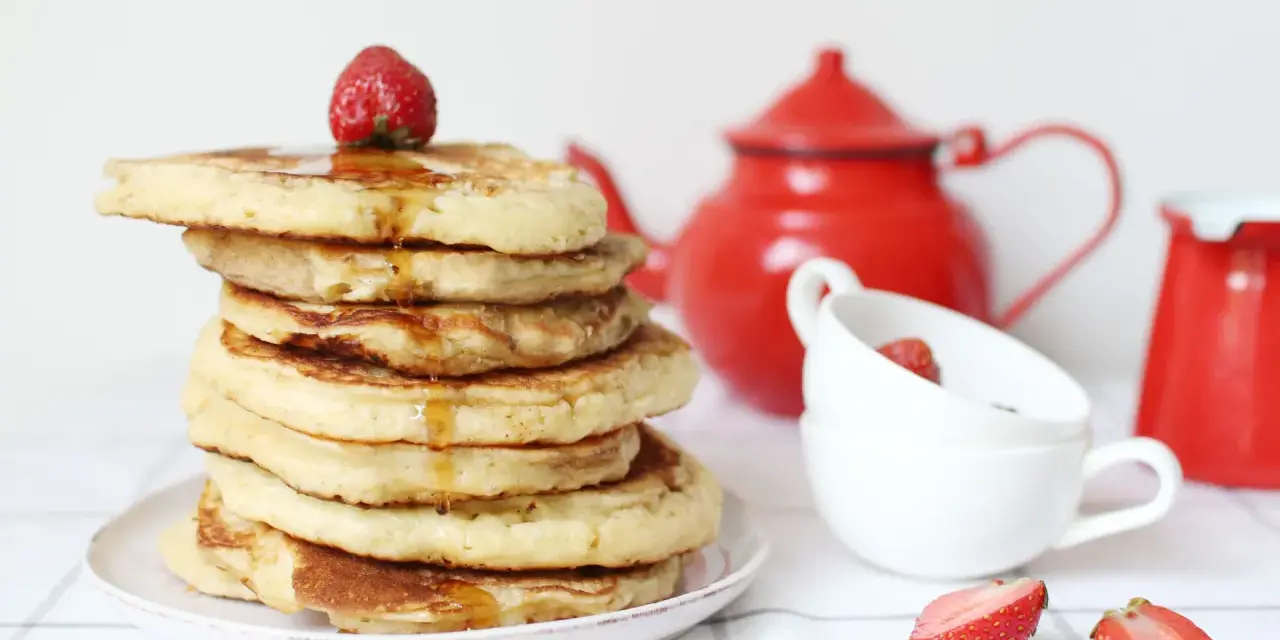 Pile de pancake sans gluten, nappés de sirop et garnis d'une fraise. Une théière rouge et des tasses complètent la scène.