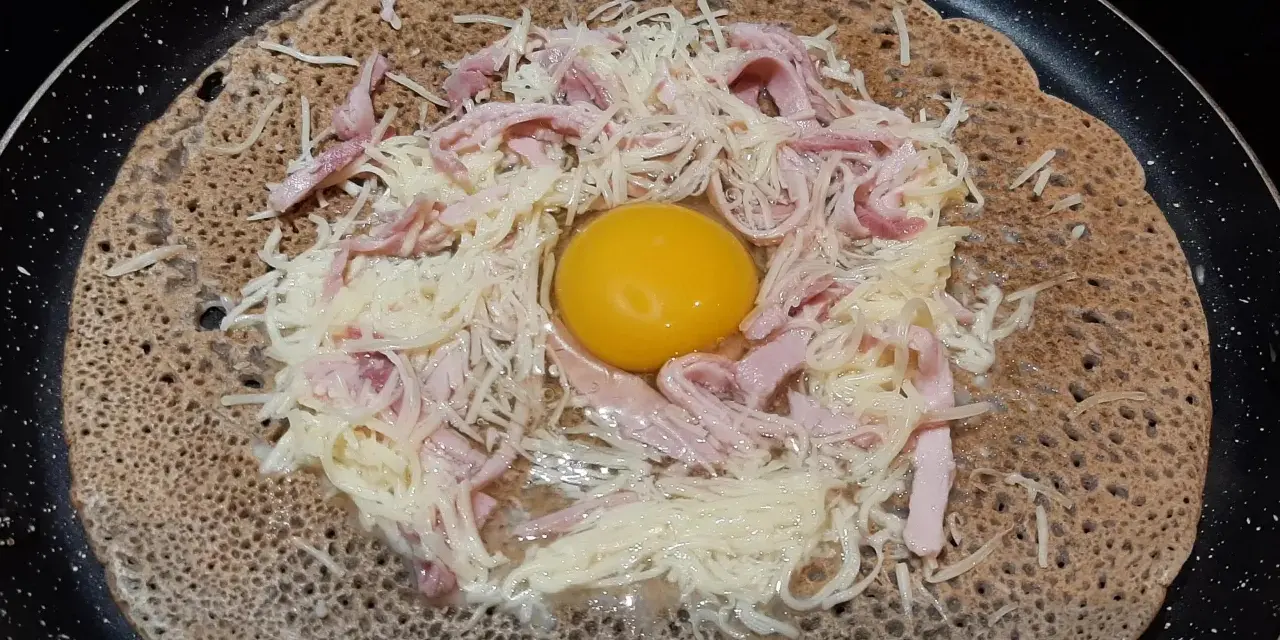 Une galette sarrasin garnie d'un œuf, de jambon et de fromage râpé, prête à être cuite dans une poêle.