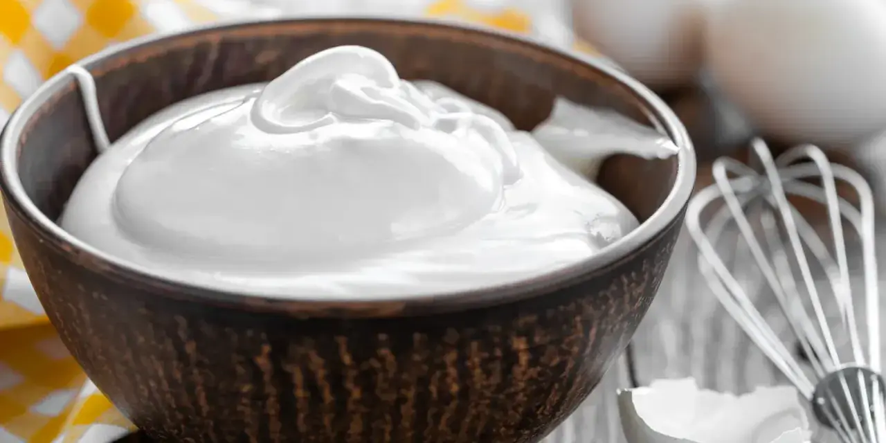 Un bol rempli de crème fouettée, blanc en neige, prêt à être utilisé pour une pâtisserie. Des œufs et un fouet sont visibles à proximité.