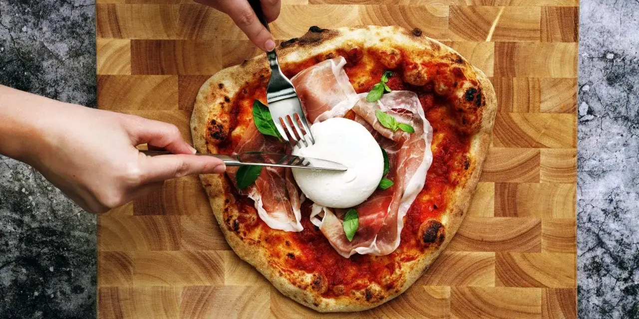 Une pizza burrata avec du prosciutto et du basilic, prête à être dégustée. Des mains coupent la burrata crémeuse.