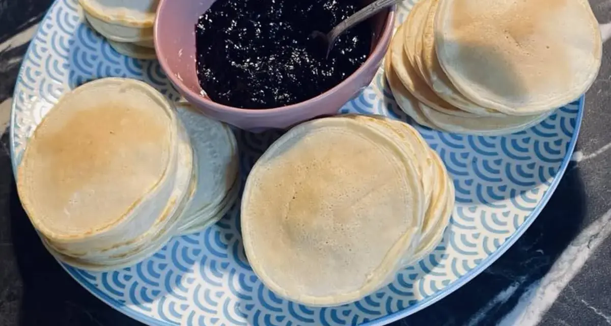 Une assiette bleue et blanche présente des piles de poffertjes dorés, accompagnés d'un bol de confiture de myrtilles. Parfait pour une recette de poffertjes !