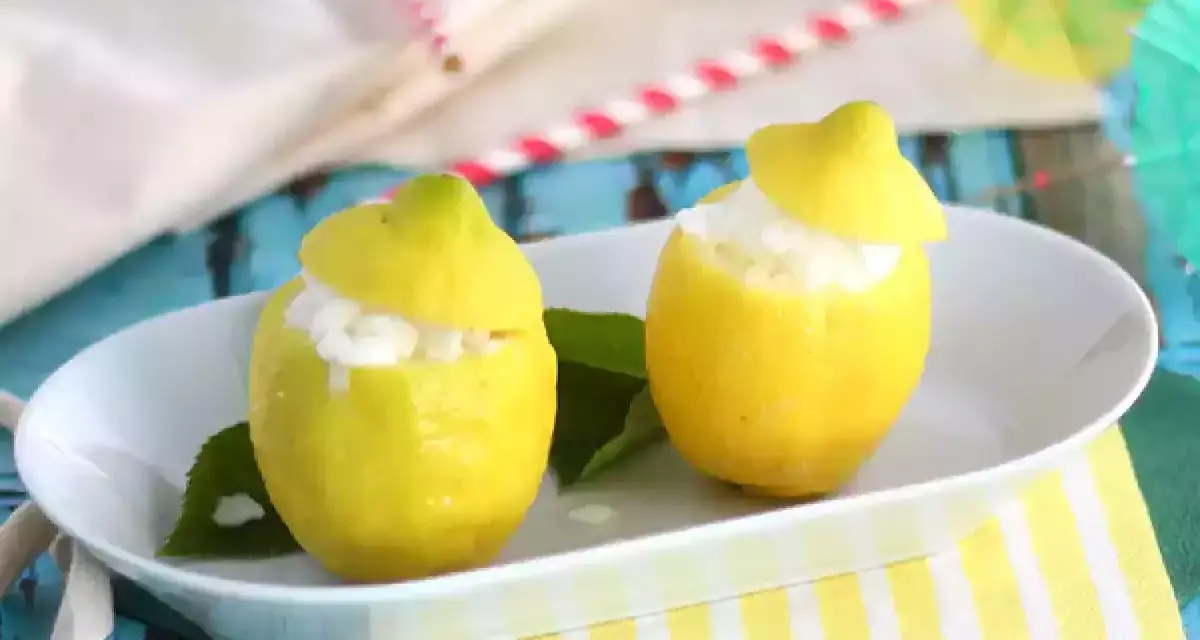 Deux citrons évidés remplis de sorbet citron, servis sur une assiette blanche, avec des feuilles vertes et des pailles colorées en arrière-plan.