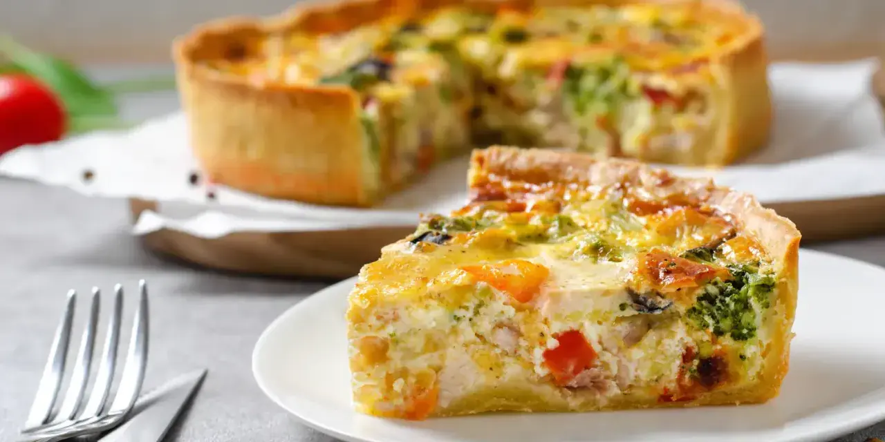 Une part de quiche savoureuse avec une pâte à tarte maison dorée, garnie de légumes et de poulet. Le reste de la tarte est flou en arrière-plan.