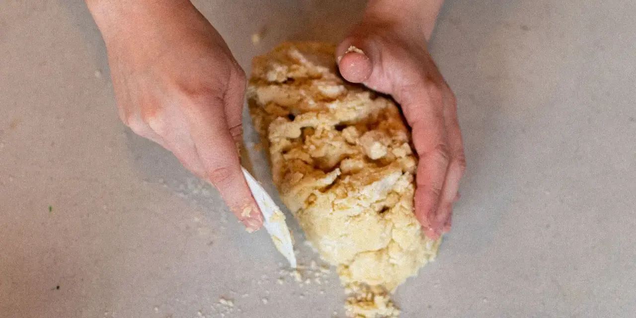 Mains travaillant une pâte sablée, la façonnant avec une spatule sur un plan de travail.