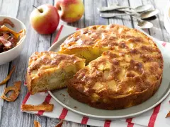 Un délicieux gâteau aux pommes, doré et moelleux, avec des morceaux de fruits visibles. Des pommes fraîches et des chips de pommes complètent la scène.