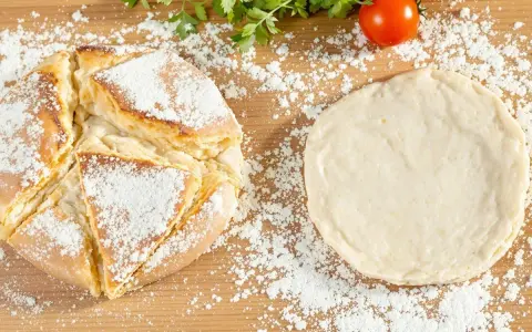 Une recette de pain maison : une miche dorée saupoudrée de farine à côté d'une pâte à pain crue, avec des tomates et du persil en arrière-plan.