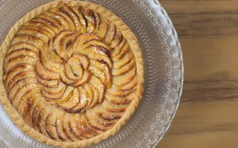 Un magnifique appareil tarte aux pommes, aux pommes caramélisées disposées en spirale sur une pâte dorée, sur un plat en verre décoré.