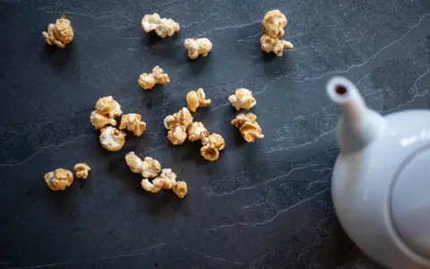 Pop corn caramel éparpillé sur une surface sombre, avec une théière blanche en arrière-plan.