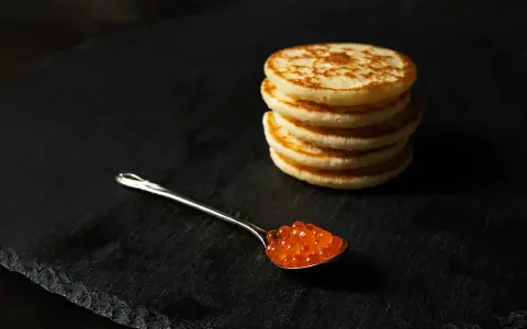 Une cuillère de caviar rouge sur une assiette noire, à côté d'une pile de blinis dorés. Parfait pour une recette blinis gourmande.