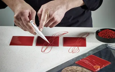Mains d'un pâtissier créant un décor royal chocolat sur des carrés rouges brillants.