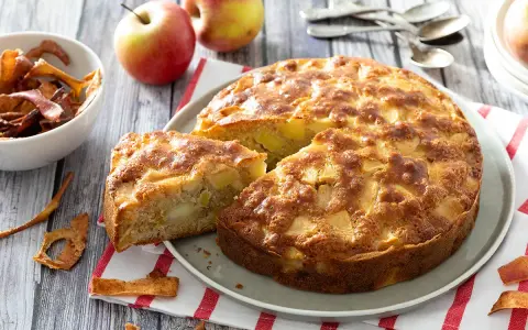 Un délicieux gâteau aux pommes, doré et moelleux, avec des morceaux de fruits visibles. Des pommes fraîches et des chips de pommes complètent la scène.