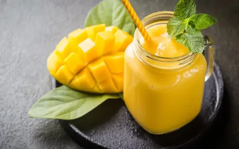 Un smoothie mangue onctueux dans un pot en verre, garni de menthe fraîche, à côté d'une moitié de mangue coupée en dés.