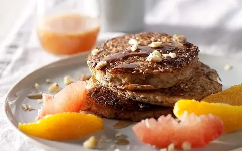 Pile de pancakes muesli, nappés de sirop et garnis de noix, accompagnés de quartiers d'agrumes.