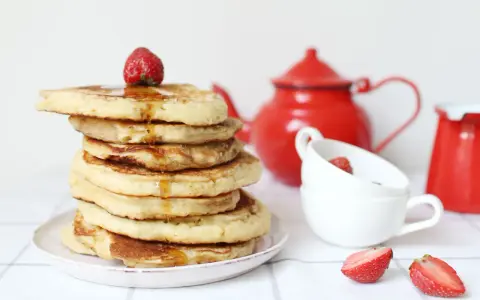 Pile de pancake sans gluten, nappés de sirop et garnis d'une fraise. Une théière rouge et des tasses complètent la scène.
