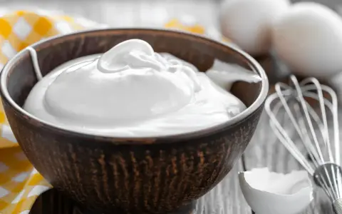 Un bol rempli de crème fouettée, blanc en neige, prêt à être utilisé pour une pâtisserie. Des œufs et un fouet sont visibles à proximité.