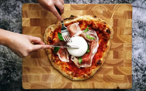 Une pizza burrata avec du prosciutto et du basilic, prête à être dégustée. Des mains coupent la burrata crémeuse.