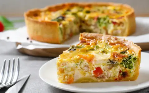 Une part de quiche savoureuse avec une pâte à tarte maison dorée, garnie de légumes et de poulet. Le reste de la tarte est flou en arrière-plan.