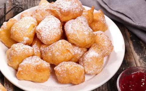 Une assiette de beignets banane saupoudrés de sucre glace, prêts à être dégustés avec une confiture de fruits rouges.