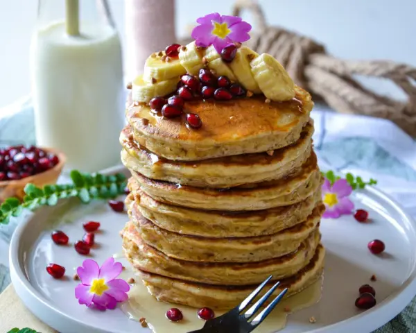 Pile de pancake banane avoine, garnie de fruits rouges, de rondelles de banane et de fleurs roses. Un délice matinal !