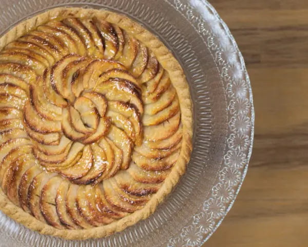 Un magnifique appareil tarte aux pommes, aux pommes caramélisées disposées en spirale sur une pâte dorée, sur un plat en verre décoré.