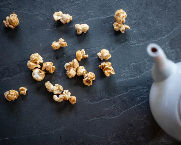 Pop corn caramel éparpillé sur une surface sombre, avec une théière blanche en arrière-plan.