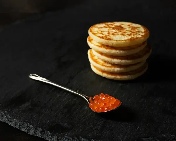 Une cuillère de caviar rouge sur une assiette noire, à côté d'une pile de blinis dorés. Parfait pour une recette blinis gourmande.